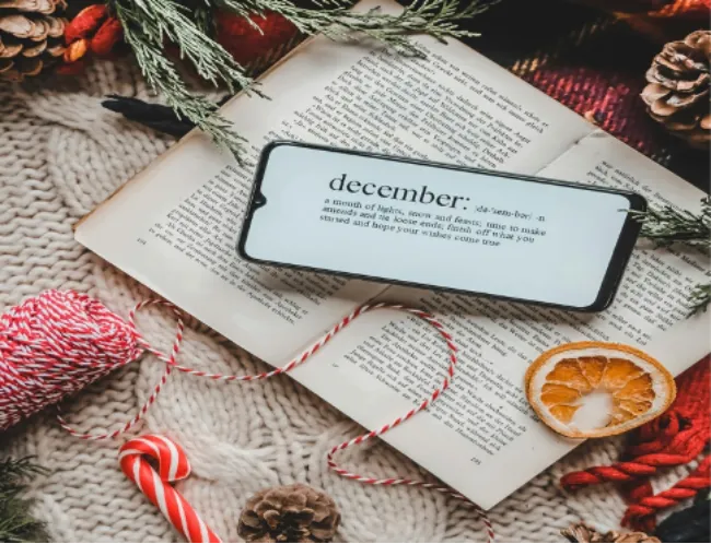a phone displaying a definition of December (that isn't fully legible) surrounded by festive December decorations such as pine cones, a candy cane, red and white string