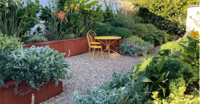 A lush garden with raised rust-colored planters filled with green foliage and flowering plants. A yellow bistro table with two matching chairs sits on a gravel pathway, surrounded by vibrant greenery.