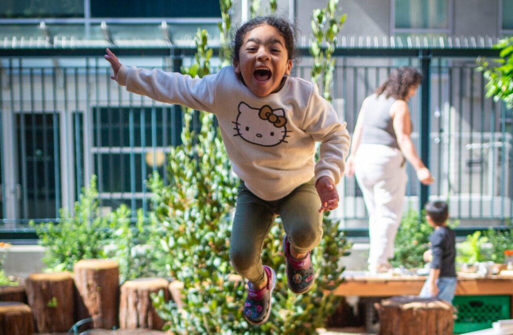 A child leaps joyfully off a carved log in a San Francisco natural playspace courtyard